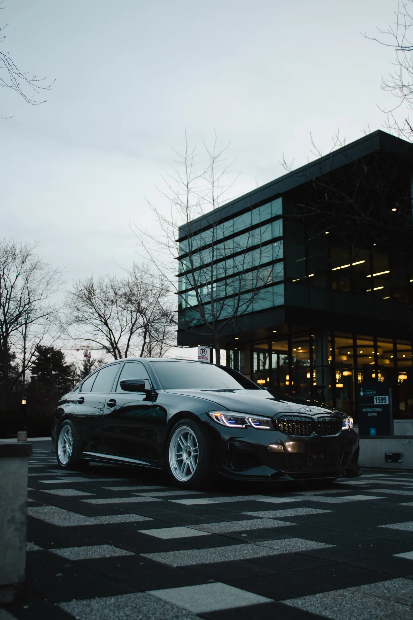 A BMW M340i on the chequered forecourt of a glass dealership on an overcast morning.