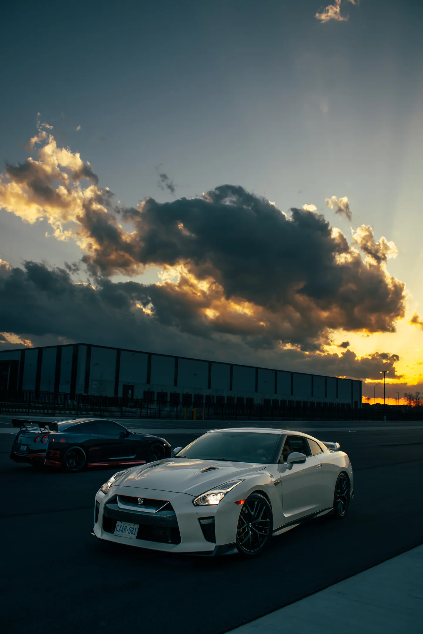 A silver Nissan GT-R in front, a black GT-R behind, under a tower of gold-lit cloud at sunset.